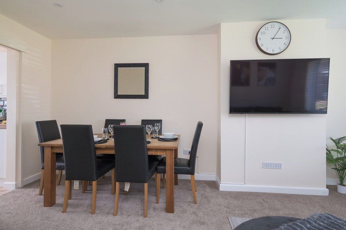 A dining area with a wooden table, black chairs, a clock, and a flat-screen TV mounted on the wall.