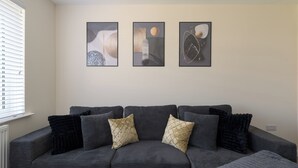 A dark grey sofa with patterned cushions, a window with blinds, and three abstract wall art pieces. - Contractor Stay | Parking & Fast Wi-Fi | Sleeps 4 (Barton Seagrave)