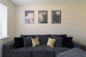 Ein dunkelgraues Sofa mit gemusterten Kissen, ein Fenster mit Jalousien und drei abstrakte Wandbilder.