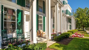Eine Reihe weißer Gebäude im Kolonialstil mit grünen Fensterläden, eine Veranda mit Stühlen und ein gepflegter Garten mit Blumen und Sträuchern.