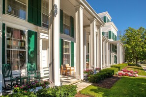 Una hilera de edificios blancos de estilo colonial, con contraventanas verdes, un porche cubierto con sillas y un jardín cuidado con flores y arbustos.