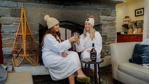 Two people in white robes sitting on a bench, holding glasses and a bottle of wine, with a stone fireplace and wicker chairs in the background. - Equinox Getaway | Queen Guest Room w/ Sauna & Spa (Manchester)