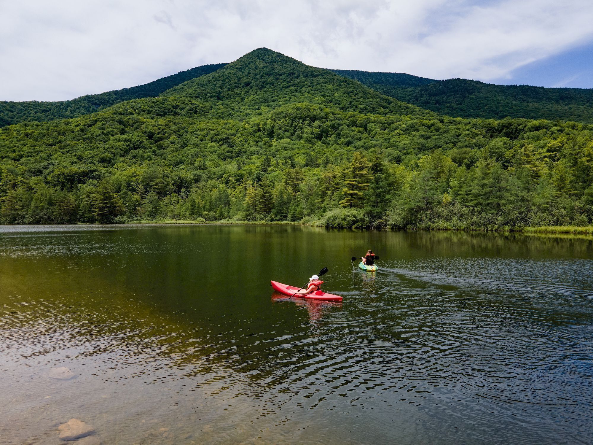 A calm lake with kayaks, surrounded by dense forest and a mountain in the background.