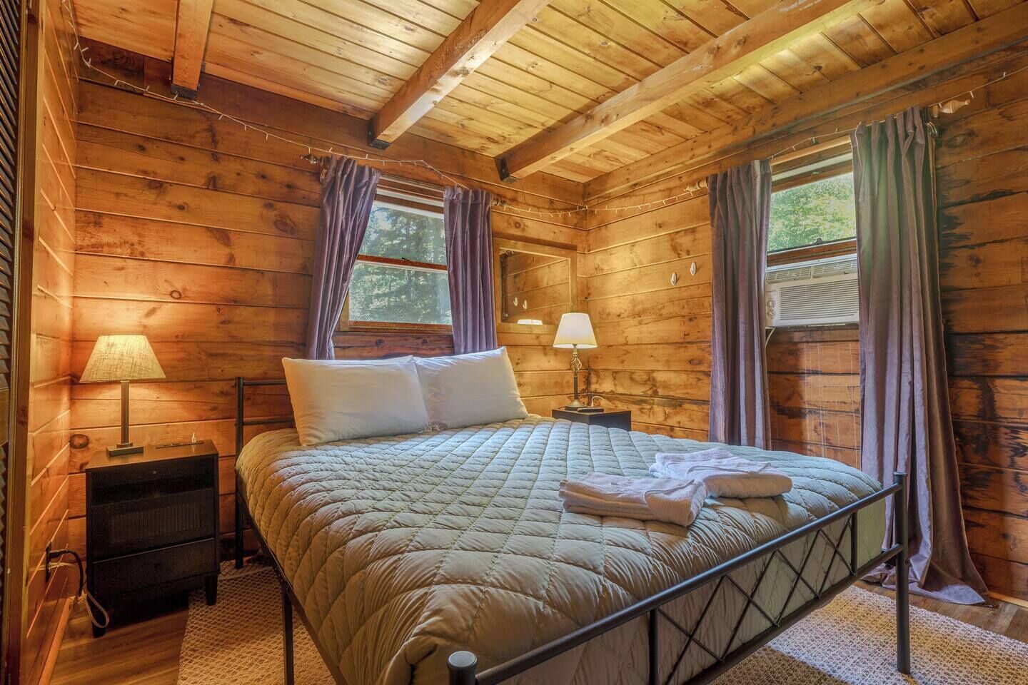 A wooden cabin bedroom with a bed, two bedside tables, and a window with curtains.