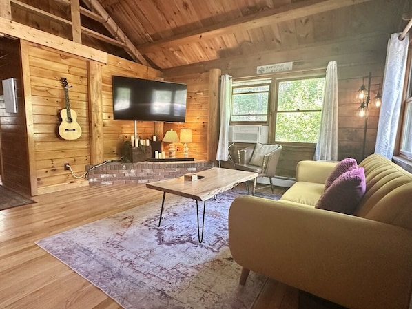 Ein Holzblockhaus-Innenraum mit Fernseher, Gitarre, Kamin, Sofa und Teppich.