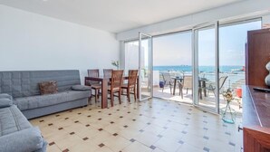 A living room with a sofa, dining table, chairs, and a view of the sea through large glass doors. - Oceanfront House, BBQ, Central Location, Easy Parking, Wi-Fi, Very Spacious (Roses)