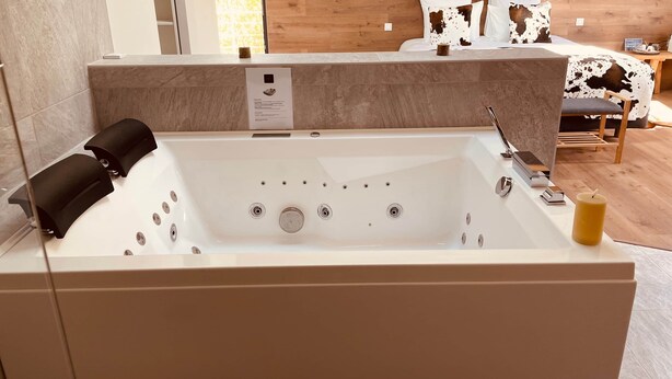 A modern bathroom with a large bathtub, a wooden wall, and a bed with a cow-patterned pillow. - Domaine Coul'Eau - Nordic Theme (Eaunes)