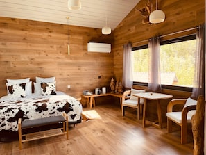 Una habitación tipo cabaña de madera con una cama, una mesita, dos sillas y un ventanal con cortinas.