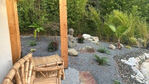 Una terraza de madera con una estatua de Buda, rodeada por un camino de grava, rocas y varias plantas.