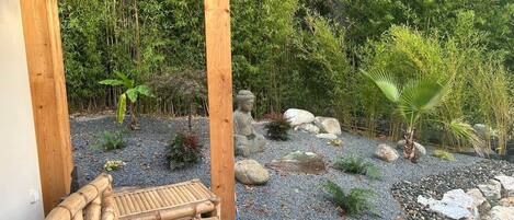 Una terraza de madera con una estatua de Buda, rodeada por un camino de grava, rocas y varias plantas.