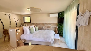 Une chambre avec un mur en pierre, un lit recouvert de linge de lit blanc, une petite table, une chaise et une fenêtre donnant sur un paysage verdoyant.