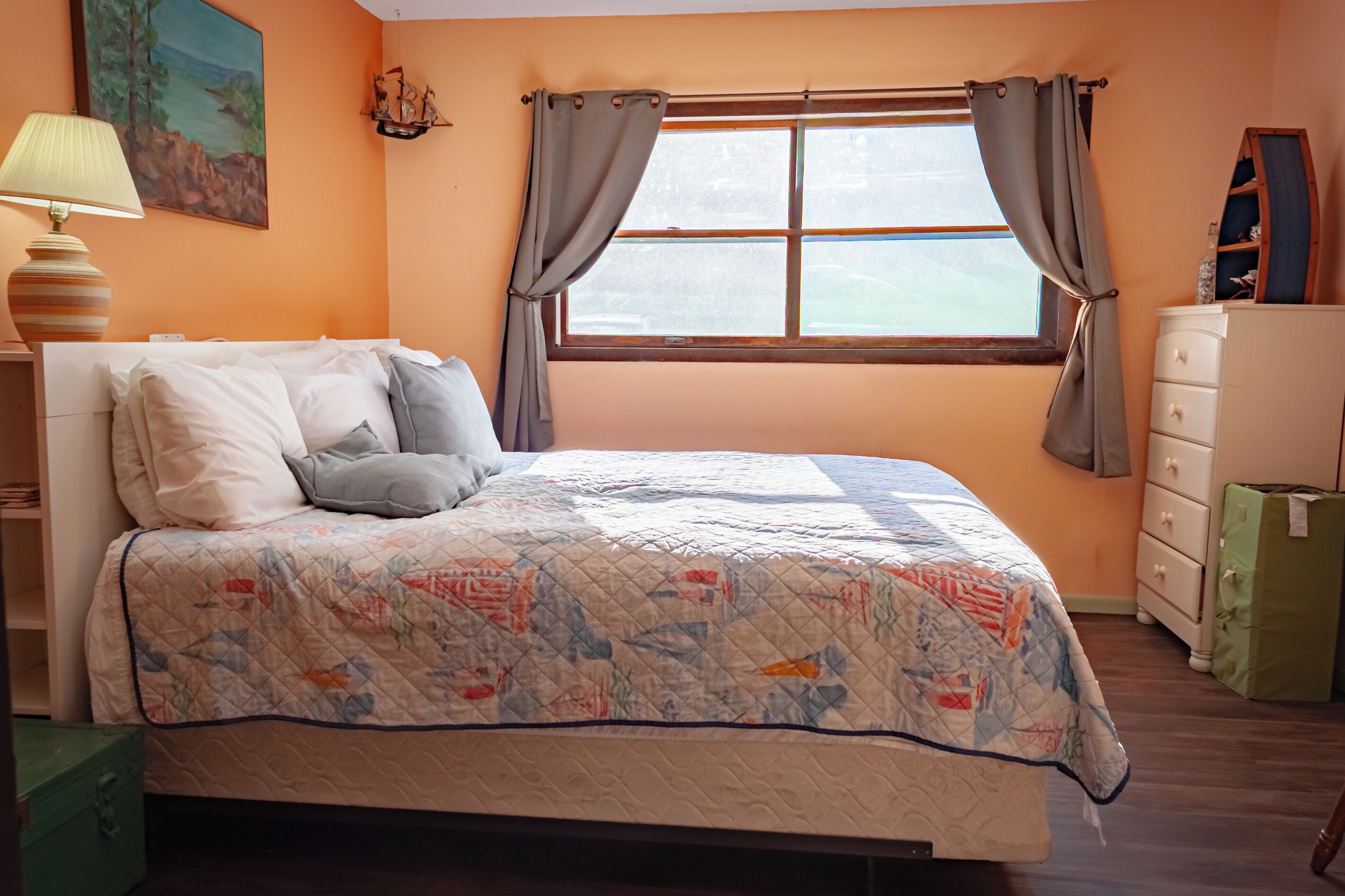 A bedroom with a bed, a dresser, a window with curtains, a lamp, and a painting on the wall.