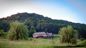 A two-story house with a balcony, surrounded by a grassy field and trees, with a forested hill in the background.