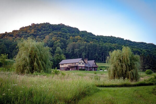 A two-story house with a balcony, surrounded by a grassy field and trees, with a forested hill in the background.