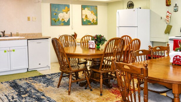 A dining area with a wooden table and chairs, a kitchen with white cabinets, and two paintings on the wall.