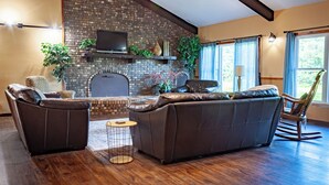 A living room with a brick fireplace, leather sofas, a television, and a large window with curtains.