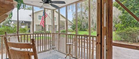 A covered porch with a ceiling fan, a wooden chair, a small table, and a view of a house with an American flag.