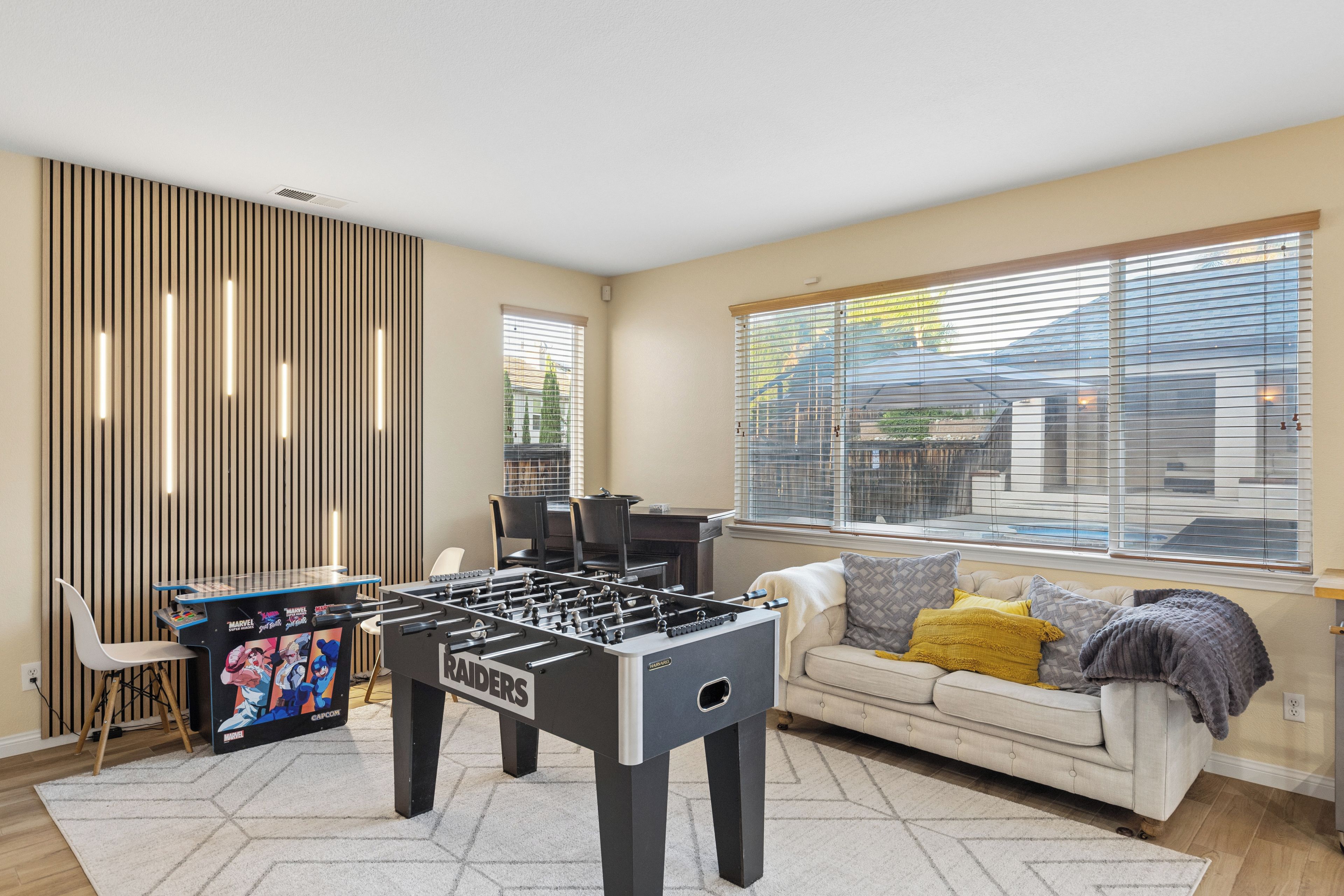 A room with a foosball table, a sofa, a window with blinds, and a wall with vertical slats.