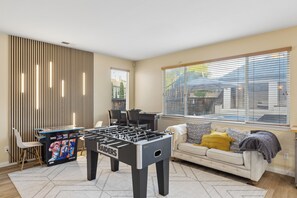 Una habitación con mesa de fútbol, sofá, ventana con persianas y una pared con listones verticales.