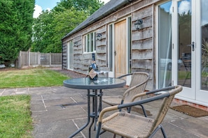 A patio with a round table and wicker chairs, a bottle of Prosecco, and a wooden building with a glass door. - Charming Countryside Barn | Rural Shropshire - Pass the Keys (Newport)