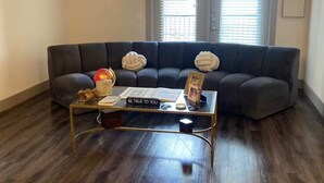 Ein Wohnzimmer mit einem dunkelgrauen Sofa, einem Glastisch mit einem motivierenden Spruch, einem Deckenventilator und einem Fenster mit Jalousien.