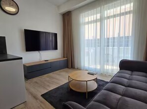 Un salon moderne avec un tĂ©lĂ©viseur Ă Ă©cran plat, un canapĂ©, une table basse et une grande fenĂȘtre avec des rideaux.