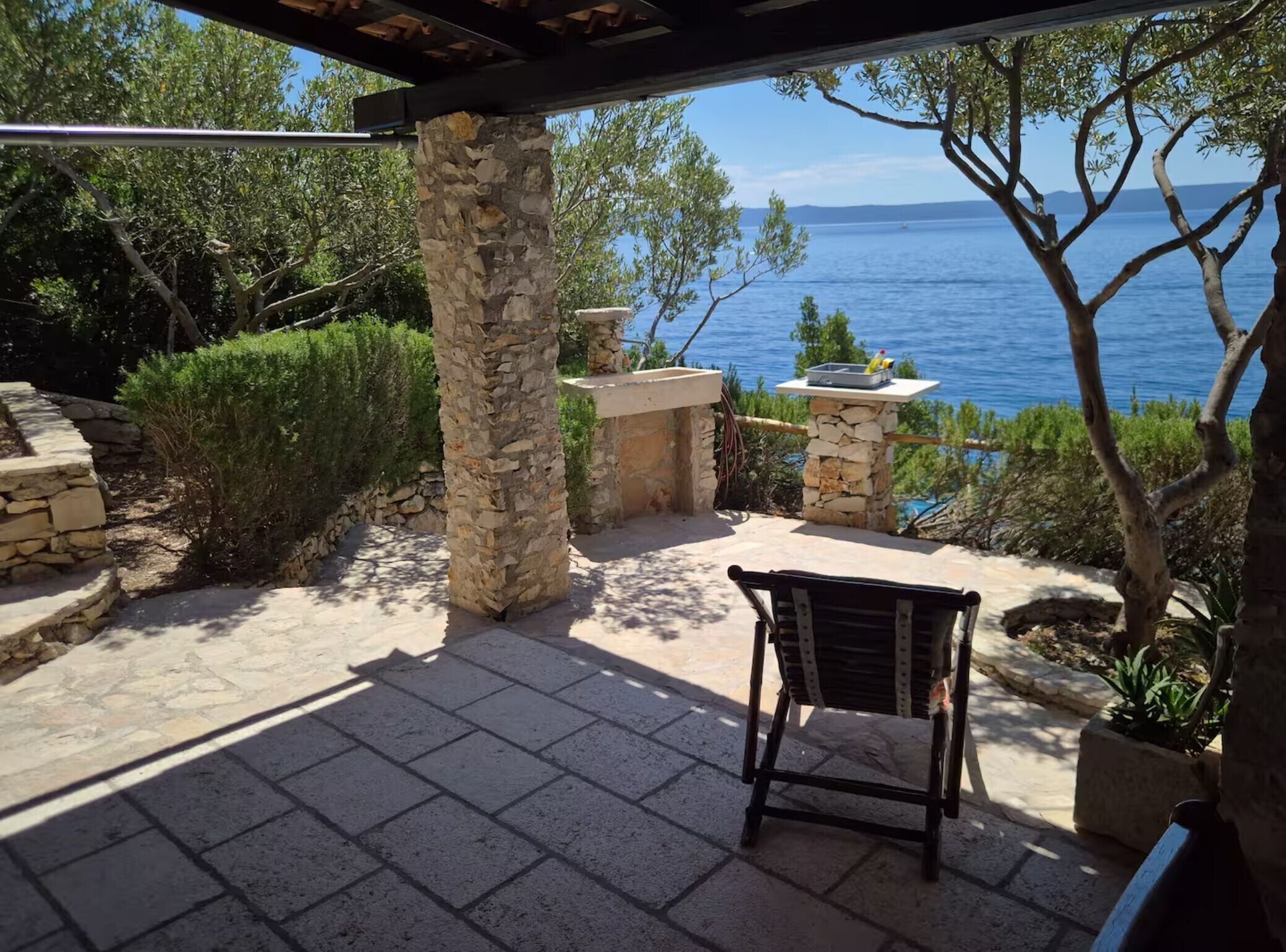 A stone pillar, a stone table, and a chair in a patio area with a view of the sea.