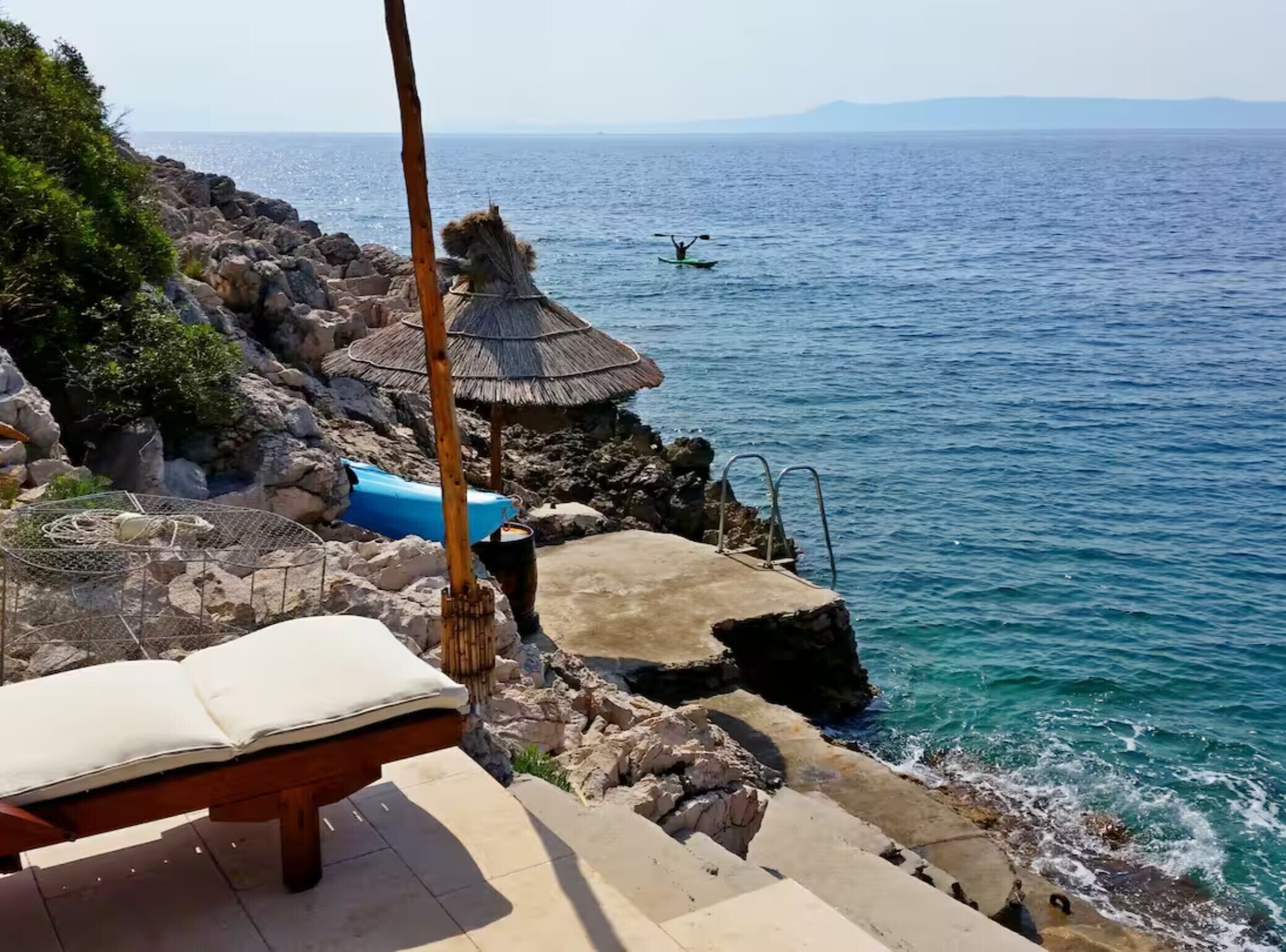 A coastal view with a rocky shoreline, a thatched-roof structure, a kayak in the water, and a lounge chair.