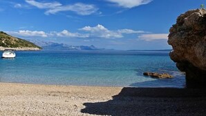 En strand med småsten, klart vatten, en båt och berg i fjärran.