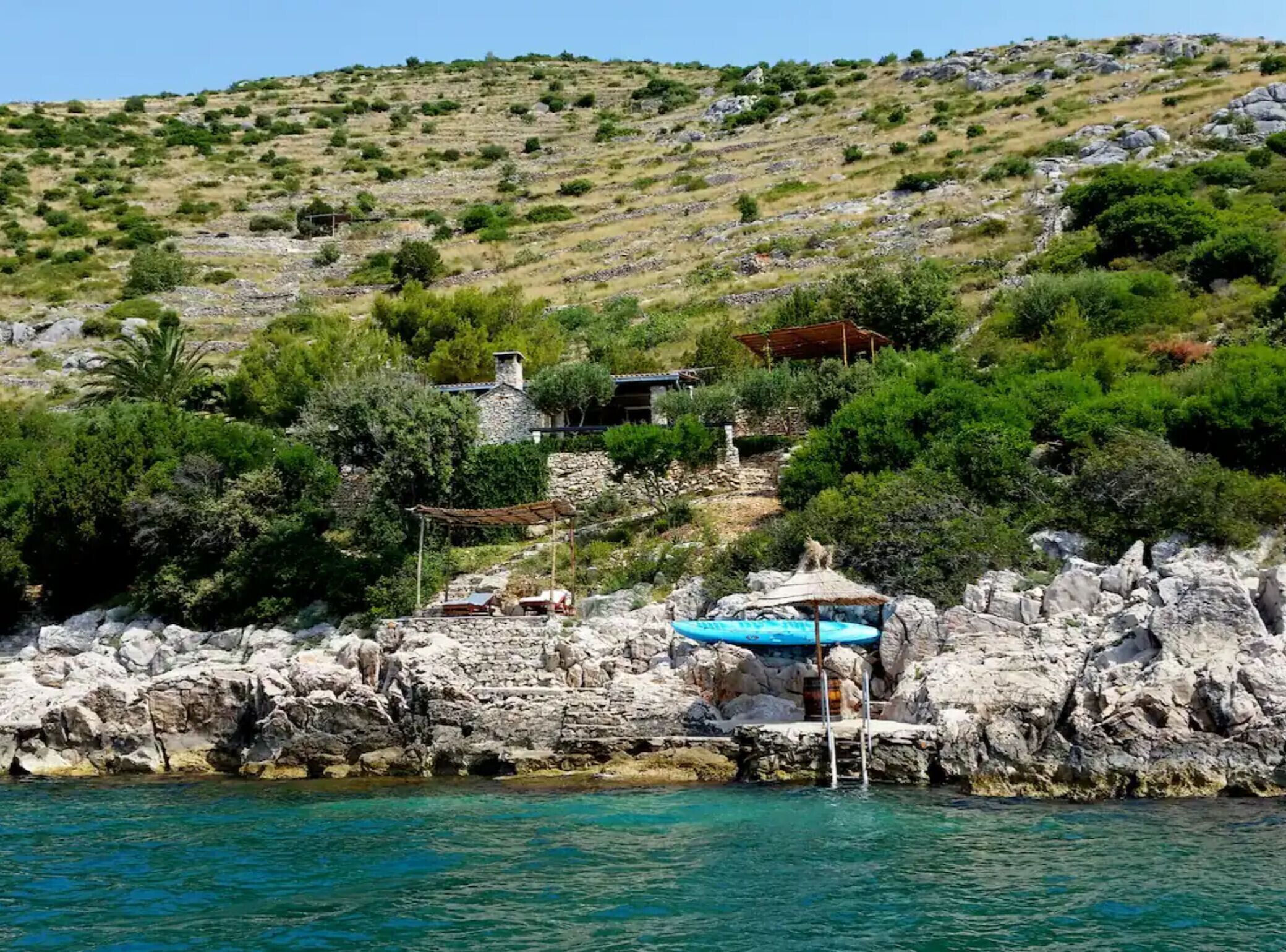 Une propriété située sur la côte, avec un bâtiment en pierre, une petite piscine recouverte d’une bâche bleue et un rivage rocheux.