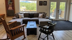 A living room with a sectional sofa, a wooden rocking chair, a small table, and a view of a deck with outdoor furniture. - Unique Container home. 10 min from town. Stunning Mountain Views! (Golden)
