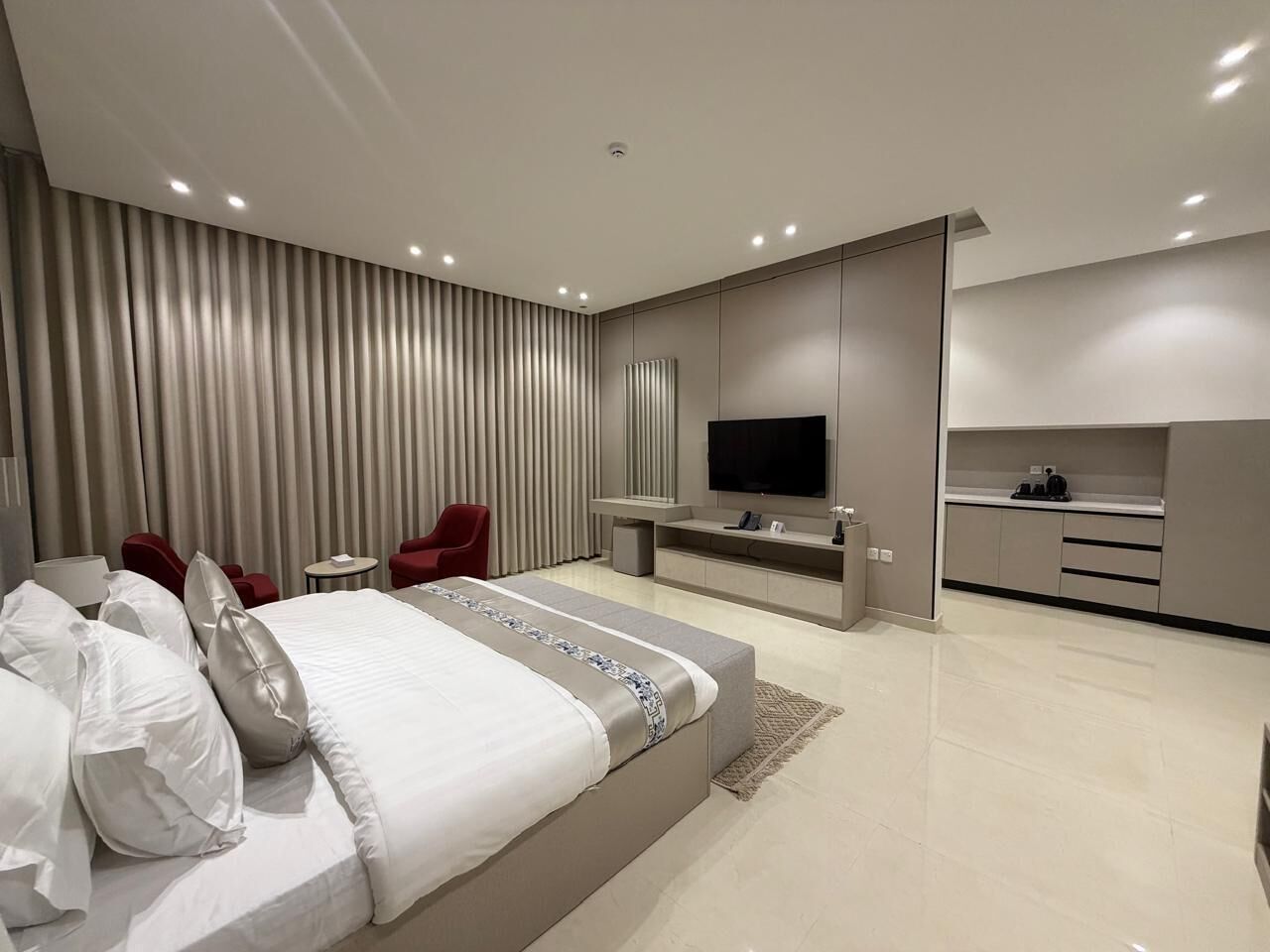 A modern hotel room with a large bed, a TV, a seating area with red chairs, and a kitchenette.