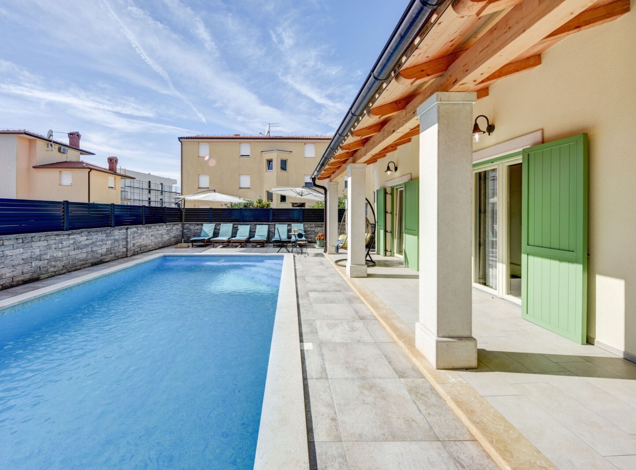 A swimming pool with lounge chairs, a covered patio, and a building with green shutters.