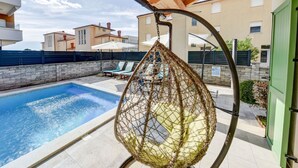 Un espace piscine agrémenté d’un fauteuil suspendu, de chaises longues et offrant une vue sur les bâtiments en arrière-plan.