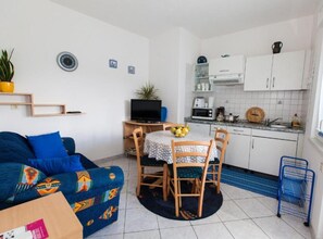 Ein gemütlicher Wohnraum mit einer Couch, einem Esstisch mit Stühlen, einem Fernseher, einer Küchenzeile mit Schränken und Geräten sowie einer kleinen Topfpflanze.