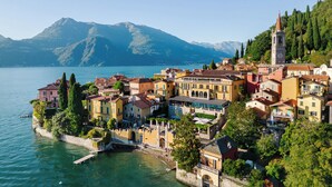 A coastal town with buildings along the water, a prominent tower, and mountains in the background. - Frenk House above Varenna (Lombardia)