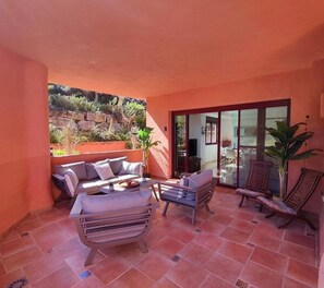 Une terrasse ensoleillée avec un canapé, des fauteuils et une table, une porte-fenêtre coulissante donnant sur un coin repas, et une vue sur une colline rocheuse.