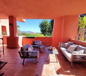 Um pátio externo com sofá, cadeiras e uma mesa, cercado por paredes alaranjadas e vista para áreas verdes e montanhas.
