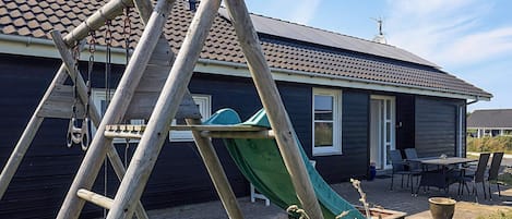 Une maison avec un toboggan vert, une échelle en bois et une terrasse dotée de mobilier d’extérieur.
