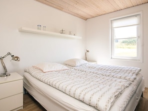 A bedroom with a bed, bedside tables, a lamp, a window, and a shelf with decorative items. - 5 Star Holiday Home in Lokken (Lokken)