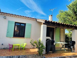 Eine Terrasse mit Tisch und Stühlen, ein kleiner Garten und ein Gebäude mit einem Ziegeldach und Fensterläden.