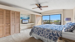 Un dormitorio con cama, ventilador de techo, televisor en un estante, ventana con vistas y balcón.