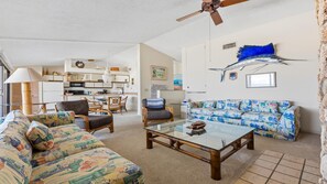 Una sala de estar con sofá, sillón y mesa de centro. Se ve una cocina al fondo con armarios y una zona de comedor.