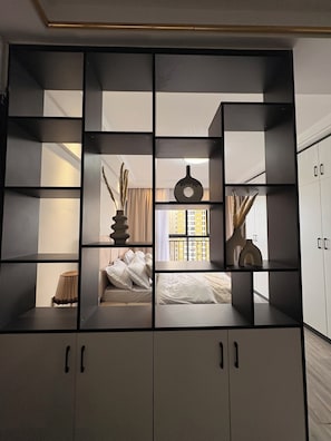 A modern room with a bed, a window with a view of buildings, and a built-in shelving unit with decorative items. - Studio in kileleshwa (Nairobi)