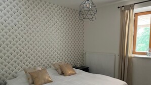 A bedroom with a bed, two pillows, a nightstand, a geometric pendant light, a window with curtains, and a radiator.