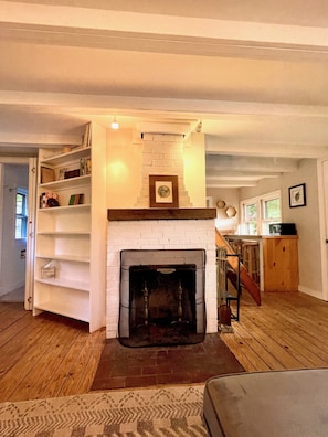 A living room with a fireplace, wooden floors, a staircase, and a kitchen area. - Charming lakefront cottage 20min to Portland (Gray)