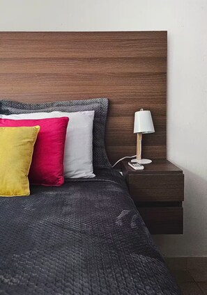 Ein Bett mit dunklem Kopfteil, ein Nachttisch mit Lampe und drei Kissen in Gelb, Rot und Weiß.