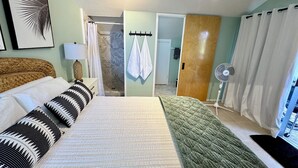 Ein Schlafzimmer mit einem Bett, Kissen, einem Nachttisch, einer Lampe, einem Ventilator, einem Spiegel und einer zur Toilette führenden Tür.
