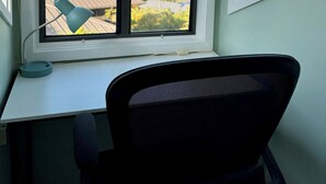 Ein Schreibtisch mit Stuhl, ein Fenster mit Blick auf ein Haus und Bäume sowie eine hängende Leuchte.