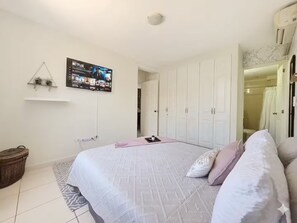 Ein Schlafzimmer mit einem Bett, einem an der Wand montierten Fernseher, einem Kleiderschrank und einem kleinen Regal mit Pflanzen.